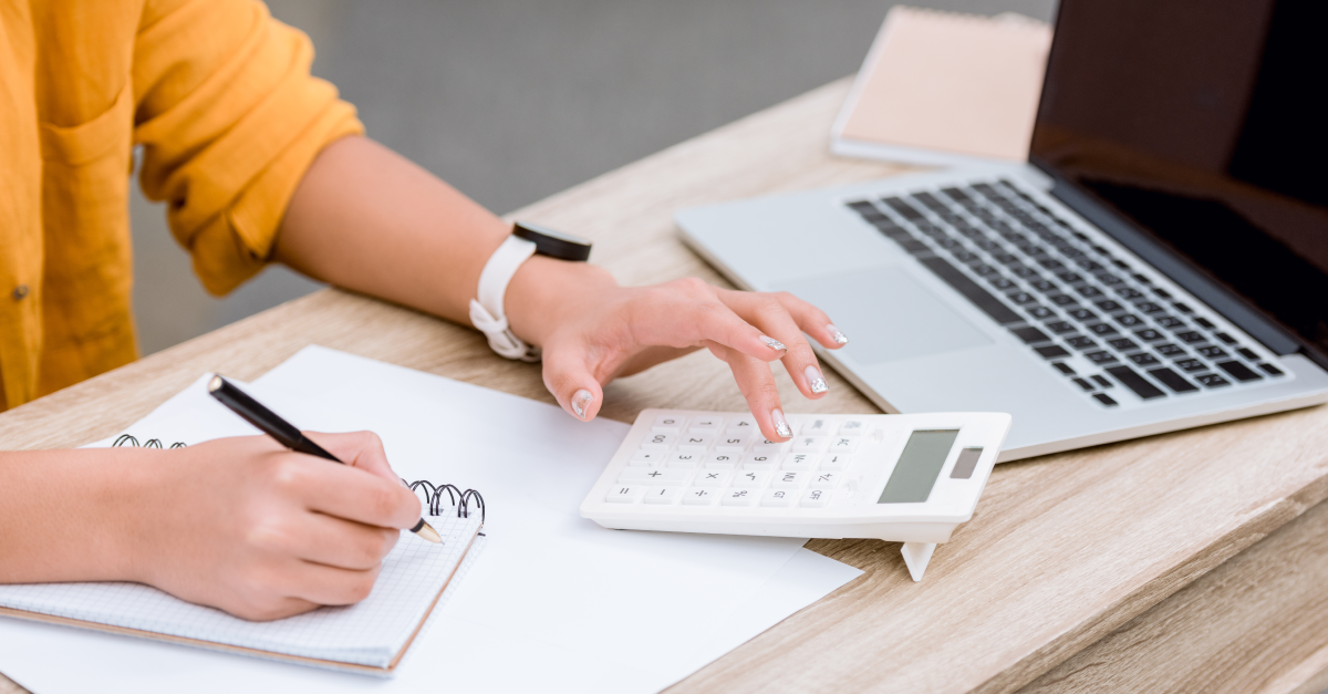 Vê-se mãos de colaboradora, que utiliza calculadora e prepara anotações